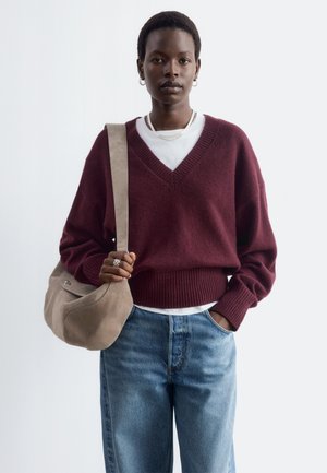 Femme portant un pull col en V bordeaux par-dessus une chemise blanche, jean bleu, boucles d'oreilles anneaux argentées, tenant un sac à épaule beige, debout sur fond blanc.