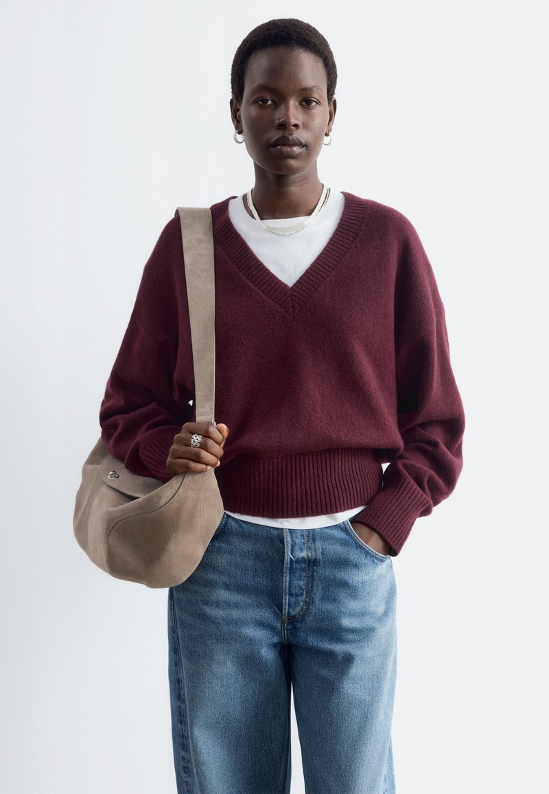 Femme portant un pull col en V bordeaux par-dessus une chemise blanche, jean bleu, boucles d'oreilles anneaux argentées, tenant un sac à épaule beige, debout sur fond blanc.