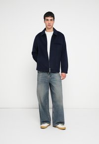 Navy krāsas jaka ar rāvējslēdzēju un apkakli, vilnas tekstūrai līdzīga, ar priekšējo kabatu. Kopā valkātas gaiši zilas plaša piegriezuma džinsa bikses un baltas sporta kurpes ar bēšām zolēm.