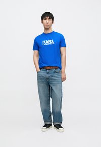 Zils kokvilnas T-krekls ar "KARL LAGERFELD" logotipu, kopā ar gaiši zilām džinsa biksēm. Modelis valkā melnas sporta kurpes un brūnu jostu.