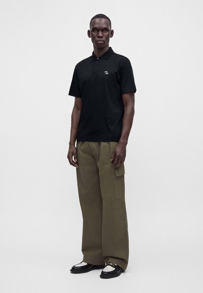 Homme debout portant un polo noir à manches courtes, un pantalon cargo vert olive et des mocassins noirs et blancs sur un fond gris uni.