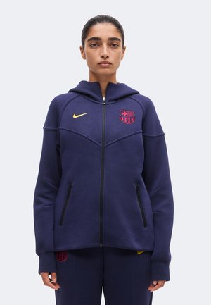 Jeune femme aux cheveux foncés tirés en arrière porte un sweat à capuche zippé bleu marine avec les logos du FC Barcelone et de Nike, debout devant un fond clair uni.