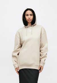 Μπεζ φούτερ με κουκούλα, εμπρόσθια τσέπη και κορδόνια, με μικρό λευκό λογότυπο στο στήθος. Χαλαρή εφαρμογή με μακριά μανίκια.