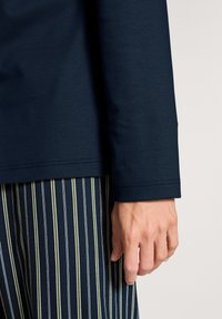 Marineblauwe longsleeve met een gladde textuur, met een net afgewerkte zoom en een close-up van een hand tegen gestreepte broeken in groen en marineblauw.