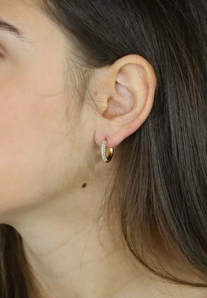 Złota kolczyk kołowy z teksturowaną powierzchnią i małymi kryształkami, prezentowany na uchu modelki z długimi, ciemnymi włosami.