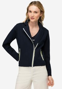 Chaqueta de marinero con un alto cuello, cierre de cremallera asimétrico y dos bolsillos frontales con cremallera. Presenta detalles de cremallera en un vibrante verde lima.