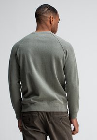 Grauer Pullover aus strukturiertem Stoff, mit Raglanärmeln und geripptem Saum, über dunklen Hosen getragen, von hinten betrachtet.