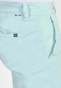 Pantalon en coton bleu clair avec une poche latérale, des accents cousus et une étiquette de logo près de la poche, affichant une texture lisse.