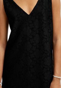 Robe en dentelle noire avec un décolleté en V, motif floral et surface texturée. Le tissu semble léger et délicat. Bracelet en argent visible.