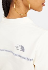 Gros plan d’une personne portant un sweat-shirt blanc North Face avec logo et texte répété au dos, et une grande boucle d’oreille dorée.