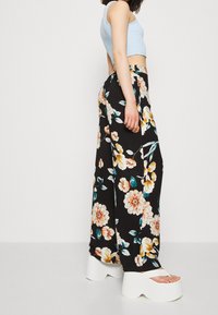 Pantalons larges à motifs floraux noirs, ornés de grandes fleurs orange et blanches, associés à des sandales à plateforme blanches et un crop top bleu clair.