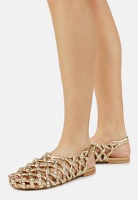 Πόδια που φορούν επίπεδα, μεταλλικά χρυσά υφαντά σανδάλια με ανοιχτό, δεμένο σχέδιο και λουράκι στον αστράγαλο σε ανοιχτό φόντο.
