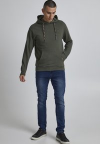Olive green hoodie with drawstrings and front pocket, paired with dark blue jeans and black sneakers. Smooth texture, casual design.