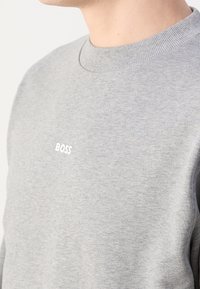 Hombre vestido con un suéter gris claro de cuello redondo, con la palabra "BOSS" bordada en blanco en el pecho.