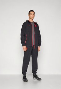 Survêtement de sport noir avec des accents rouges, comprenant une veste zippée et un pantalon fuselé. Fabriqué en matériau léger, il comprend une capuche et des poches.