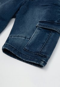 Pantaloni cargo in denim di colore blu scuro con una finitura testurizzata. Caratterizzati da una tasca laterale, orlo arrotolato e dettagli di cucitura a vista.
