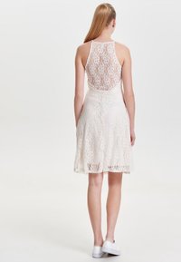 Kermarin pitsi mekko, jossa on selkäaukkoinen muotoilu, kaula-aukko ja aaltonen helma. Yhdistetty valkoisiin tennareihin rentoa tyyliä varten.