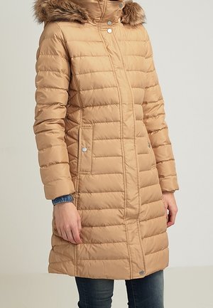 Długi beżowy pikowany zimowy płaszcz z kapturem obszytym futerkiem, zatrzaskami i bocznymi kieszeniami, noszony na niebieskich dżinsach.