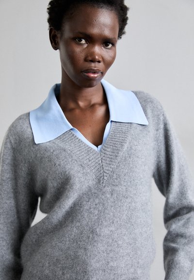 Jersey gris de punto con escote en V, combinado con una camisa de color azul claro debajo. Textura suave, corte relajado y detalles de diseño mínimos.