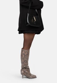 Bottes en imprimé serpent, montantes jusqu'au genou, en beige et marron, avec des bouts pointus et un talon fin, associées à un sac à main noir et une jupe noire à volants.
