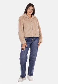 Giacca in pelliccia sintetica tan con zip e collarino, abbinata a jeans blu e scarpe da ginnastica bianche. La giacca ha una vestibilità comoda e una texture morbida.