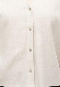 Gros plan d'une chemise en tissu texturé de couleur crème avec quatre boutons beiges fermés au centre devant.