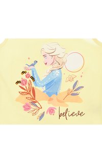 Canotta gialla con un'immagine di un personaggio dai capelli biondi e un vestito blu, che tiene in mano un piccolo uccello tra design floreali e la parola "credi".