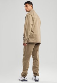 Giacca di tela beige con vestibilità rilassata, tasche laterali e pantaloni beige coordinati; abbinata a sneakers bianche con dettagli grigi.