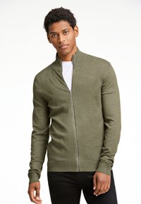 Olivgrön zip-up tröja med hög krage, figursydd design och ribbstickade muddar. Bärs över en vit T-shirt. Mjuk stickad textur.