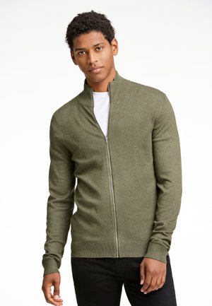 Olivgrüner Reißverschluss-Pullover mit hohem Kragen, tailliertem Design und gerippten Bündchen. Getragen über einem weißen T-Shirt. Weiche Strickstruktur.