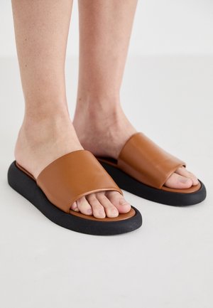 Persoon draagt bruine slippers met brede leren banden en dikke zwarte zolen op een glad, lichte ondergrond.