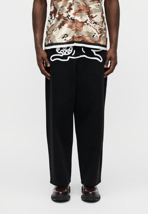 Személy fekete, bő szárú nadrágot visel, amelyen fehér grafikai minta található a derékrésznél, barna terepmintás gombos ingben és sötétbarna cipőben.