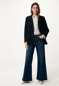 Svart dubbelknäppt blazer över en vit, strukturblus, kombinerad med vida blå denimjeans och ljusa skor.