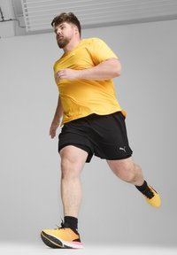 Maglietta sportiva gialla a maniche corte, pantaloni neri e scarpe da ginnastica arancioni con accenti neri; uomo che corre su uno sfondo grigio uniforme.