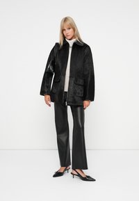 Giacca nera in pelliccia sintetica con due tasche frontali, chiusura con zip e colletto a contrasto, abbinata a pantaloni in pelle nera a vita alta e scarpe nere.