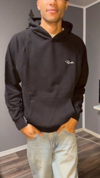 Black hoodie with a front pocket and adjustable drawstring, worn with light blue jeans. Hoodie has a small white logo on the chest.