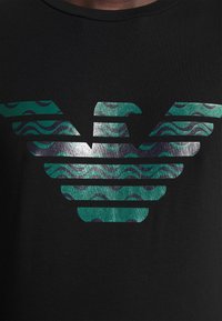 Černé tričko s logem se zelenými a tyrkysovými vlnitými vzory a metalickými akcenty. Design je symetrický a výrazně umístěný na přední straně.