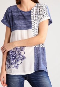 Blå och vit kortärmad topp med en blandad mönsterdesign, med ett stort blått blommotiv och texturerade tygalternativ i olika nyanser.