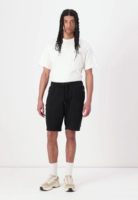 Svart bomullsshorts med elastisk midje og snøring, kombinert med en hvit t-skjorte. Modellen har på seg beige sneakers med hvite sokker.