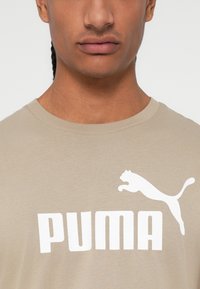 Bež pamučna majica s velikim bijelim logotipom "PUMA" i grafikom skakačkog pumaira na prednjoj strani. Kratki rukavi i okrugli izrez.