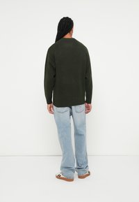 Oversized donkergroen gebreid sweater met ronde hals, geribbelde zoom, gecombineerd met lichtblauwe rechttoe rechtaan jeans en een paar sandalen met een patroon.