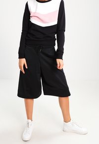 Svart sweatshirt med vit och rosa zick-zackdesign, parat med löst sittande svarta shorts och vita sneakers. Avslappnad atletisk klädsel.