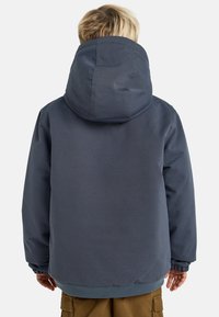 Bambino con capelli biondi che indossa una giacca con cappuccio blu scuro e pantaloni marroni, girato di spalle su uno sfondo chiaro e uniforme.