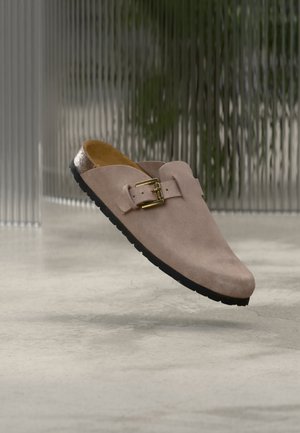 Sabot unique en daim beige avec boucle dorée et semelle noire, présenté en lévitation au-dessus d'un sol en béton lisse avec un arrière-plan flou de verre vertical.