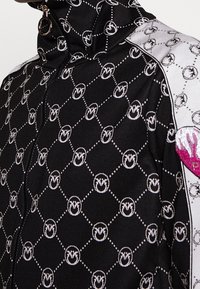 Black zip-up jacket featuring a geometric pattern with white dots and logos. Silver accents on the shoulders and a pink embroidered detail.