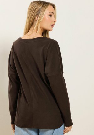 Blondynka ubrana w luźną, ciemnobrązową bluzę z długim rękawem i jasnoniebieskie dżinsy, obrócona lekko, pokazując tylny i boczny profil.