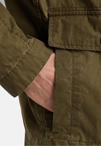 Main reposant dans la poche d'une veste vert olive, montrant la texture du tissu et les détails visibles des coutures.