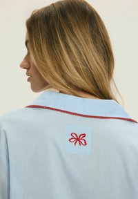 Kobieta o jasnobrązowych włosach, ubrana w jasnoniebieską koszulę z czerwonym haftowanym kokardą i czerwonym obszyciem na kołnierzu z tyłu, zwrócona w lewo.