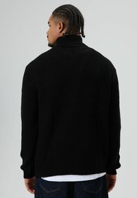 Man met gevlochten haar, gekleed in een zwarte geribbelde coltrui en donkere jeans, van achteren gezien tegen een effen grijze achtergrond.