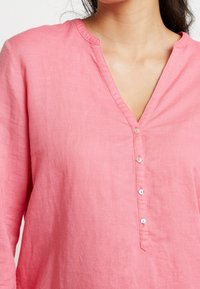 Camicia di lino rosa con scollo a V, chiusura con bottoni e maniche lunghe. Texture morbida e design casual, ideale per il clima caldo.